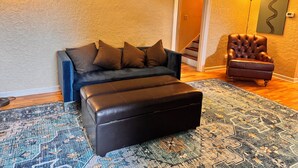 A living room with a dark sofa, a brown leather ottoman, a patterned rug, a brown armchair, and a staircase in the background.