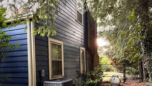 A house with a dark blue exterior, a large air conditioning unit, and a covered outdoor area with a tree and a fence.