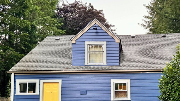 A house with a blue exterior, a yellow door, and two blue Adirondack chairs on a wooden deck.