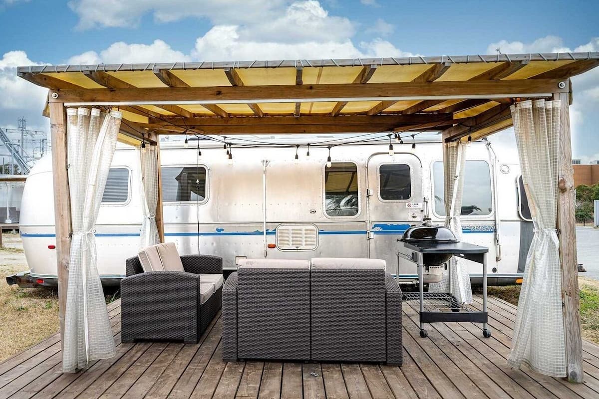 A covered outdoor seating area with wicker furniture and a grill, adjacent to a camper van.