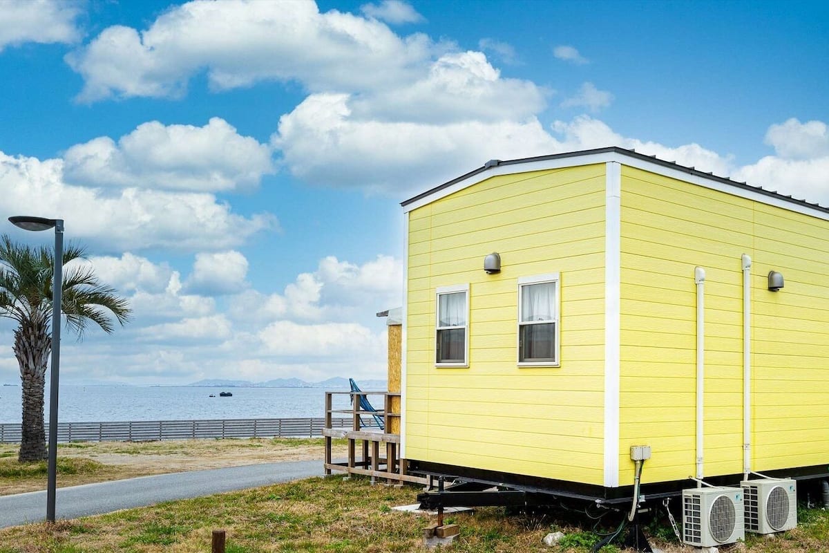 A yellow mobile home with a small deck, a palm tree, and a view of the ocean.