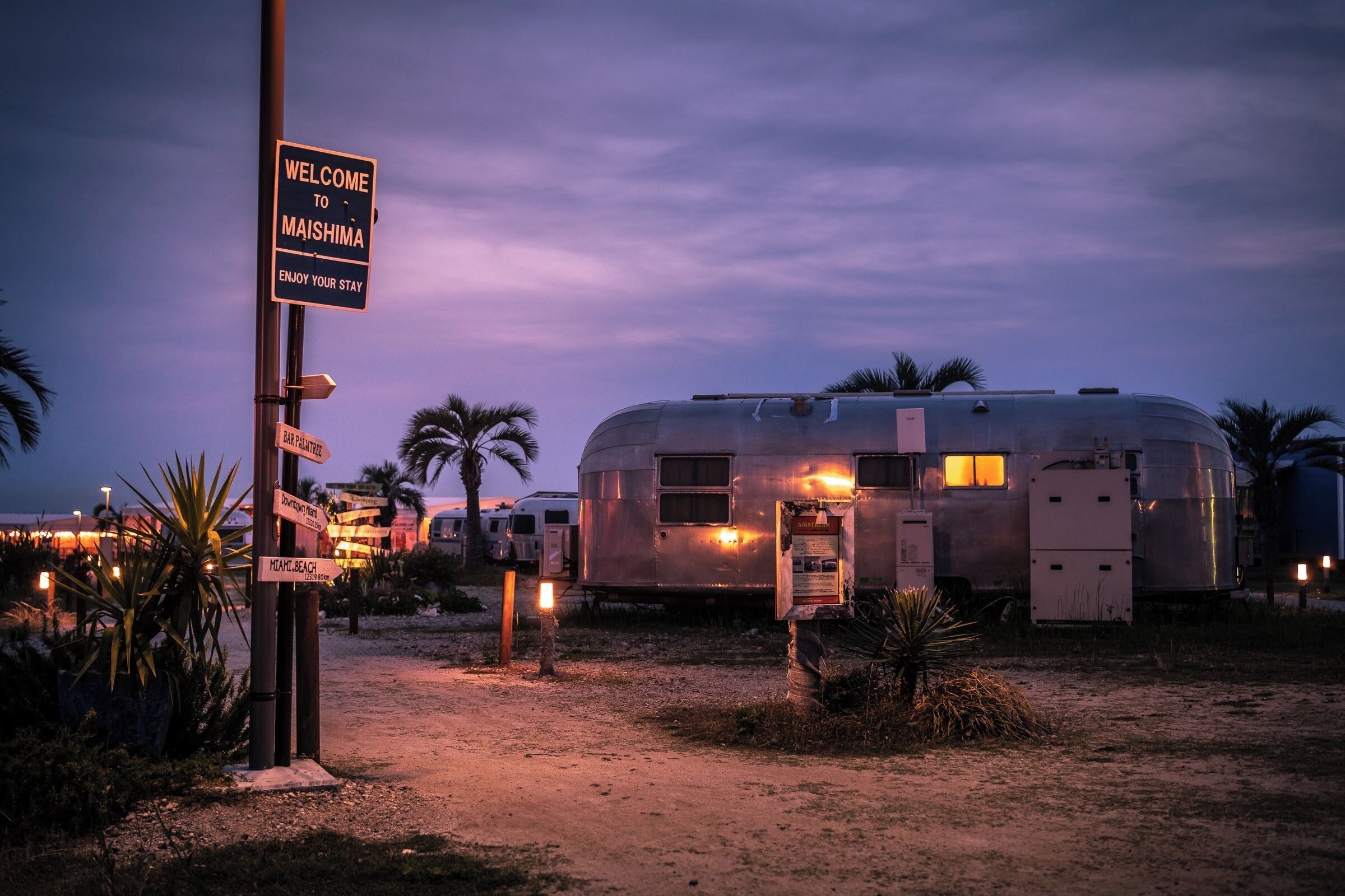 Un terrain de camping pour caravanes avec une pancarte indiquant « Bienvenue à Maishima » et des panneaux indiquant les directions vers les lieux d’intérêt à proximité.