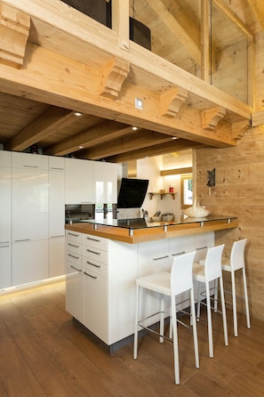 Una cocina moderna con vigas de madera, armarios blancos, una isla de vidrio y taburetes blancos.