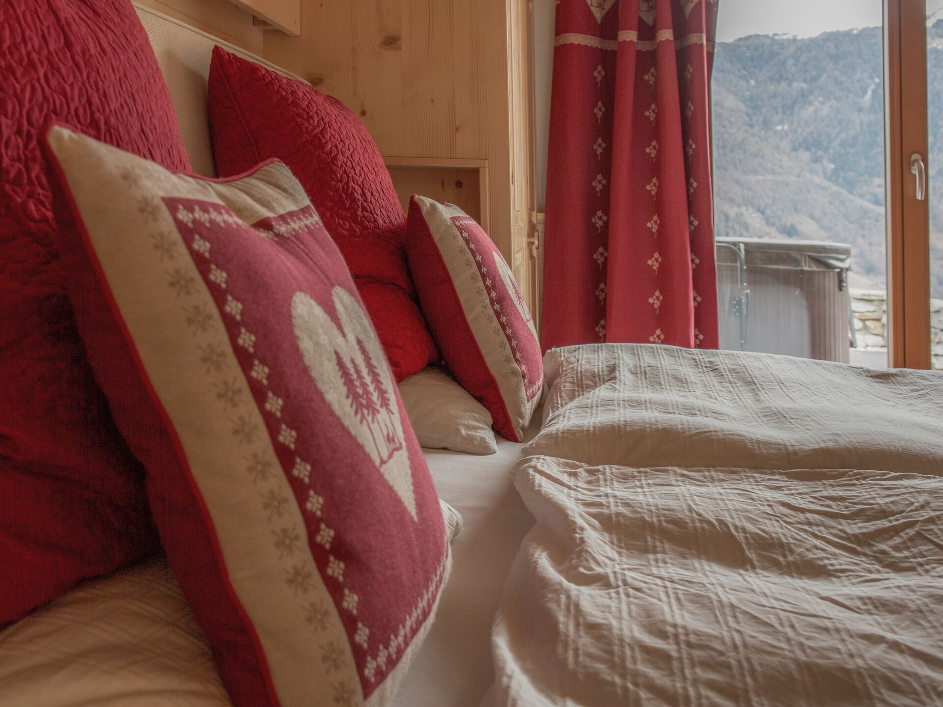 A bed with red and white pillows, a wooden headboard, and a view of mountains through a window with red curtains.