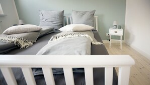 Una cama bien hecha con ropa de cama gris y almohadas blancas, una mesita de noche con una lámpara y un pequeño mueble auxiliar con un florero.