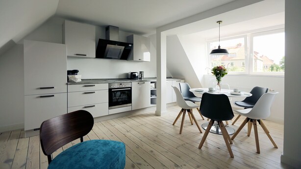 Una cocina moderna con armarios blancos, un comedor con una mesa redonda y sillas, y un ventanal que proporciona luz natural.