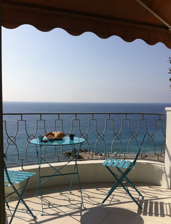 A balcony with a table and chairs, overlooking a beach with people and a clear sky.