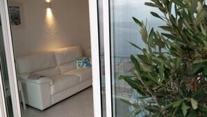 Un salon moderne avec un canapé blanc, un coussin décoratif et une porte en verre donnant sur un balcon avec vue sur la ville.