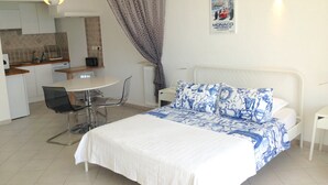 Une chambre avec un lit, une table de chevet, une chaise, une petite table, un radiateur, un rideau, une affiche au mur et, au fond, une kitchenette.