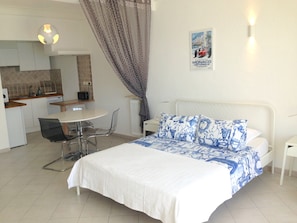 A bedroom with a bed, a bedside table, a chair, a small table, a radiator, a curtain, a poster on the wall, and a kitchenette in the background.