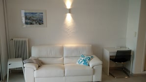 Un canapé en cuir blanc agrémenté d’un coussin à motifs bleus, une petite table d’appoint blanche, un radiateur, un tableau encadré au mur et un bureau avec une chaise.