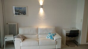 A white leather sofa with a blue patterned pillow, a small white side table, a radiator, a framed picture on the wall, and a desk with a chair.