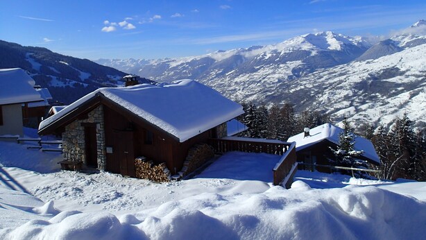 Un chalet de montagne enneigé, avec une façade en pierre et un toit en bois, entouré d’arbres et de montagnes également couverts de neige.