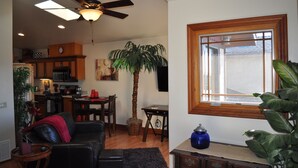A living room with a black leather sofa, a wooden coffee table, and a dining area with a wooden table and chairs. There is a kitchen in the background with wooden cabinets and a microwave. A ceiling fan is visible, and there is a large window with a wooden frame.