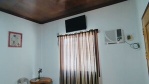 Una habitación pequeña con piso de baldosas, techo de madera, televisor montado en la pared, una ventana con cortinas, una silla, una mesita y dos botes de basura etiquetados como "MALATA" y "BILI MALATA".