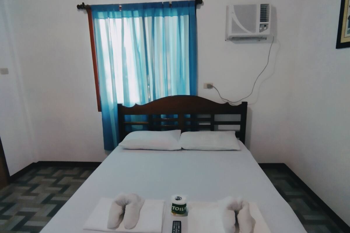 A single bed with a wooden frame, a remote control, and two pillows on a patterned floor.