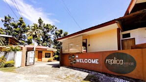Una entrada cerrada con un cartel de 'Welcome' y la marca 'Replica Manor', que conduce a un edificio con techo rojo y un área cubierta.