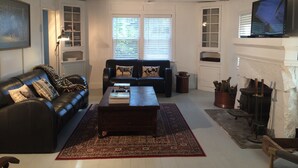 A living room with a black leather sofa, a wooden coffee table, and a red patterned rug. There is a fireplace with logs and a television mounted on the wall. - Chic Historic Cottage in the Heart of 3 Rivers-close to SNP, Hot Tub, Love dogs! (Three Rivers)