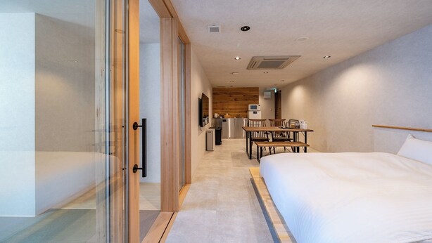 Une chambre d’hôtel moderne dotée d’un grand lit, d’un coin repas avec des tables en bois, d’un espace cuisine et d’une porte-fenêtre coulissante donnant sur un balcon.