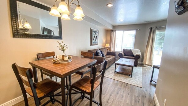 A dining area with a wooden table and chairs, a sofa, and a coffee table. There is a large mirror on the wall and a window with curtains. - 14-Plex Condo #114 Ground level, close to seasonal pool & spa (Garden City)