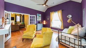 Een kamer met paarse wanden, een plafondventilator, een bed met veel kussens, een gele fauteuil en een klein tafeltje.