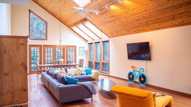 A spacious living room with a high ceiling, wooden beams, a large sofa, a yellow armchair, a dining area with a table and chairs, and a television mounted on the wall.