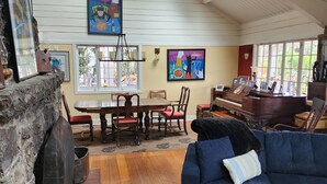 A spacious living room with a wooden floor, a blue sofa, a dining area with a table and chairs, a piano, and artwork on the walls.