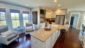 A modern kitchen with a large island, white cabinetry, and a view of the outdoors through large windows.