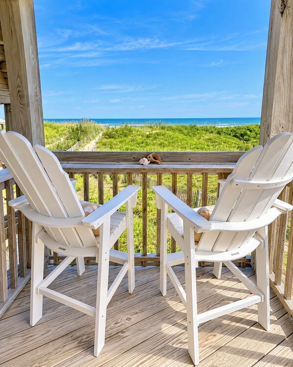 A wooden deck with two white Adirondack chairs facing a beach view.