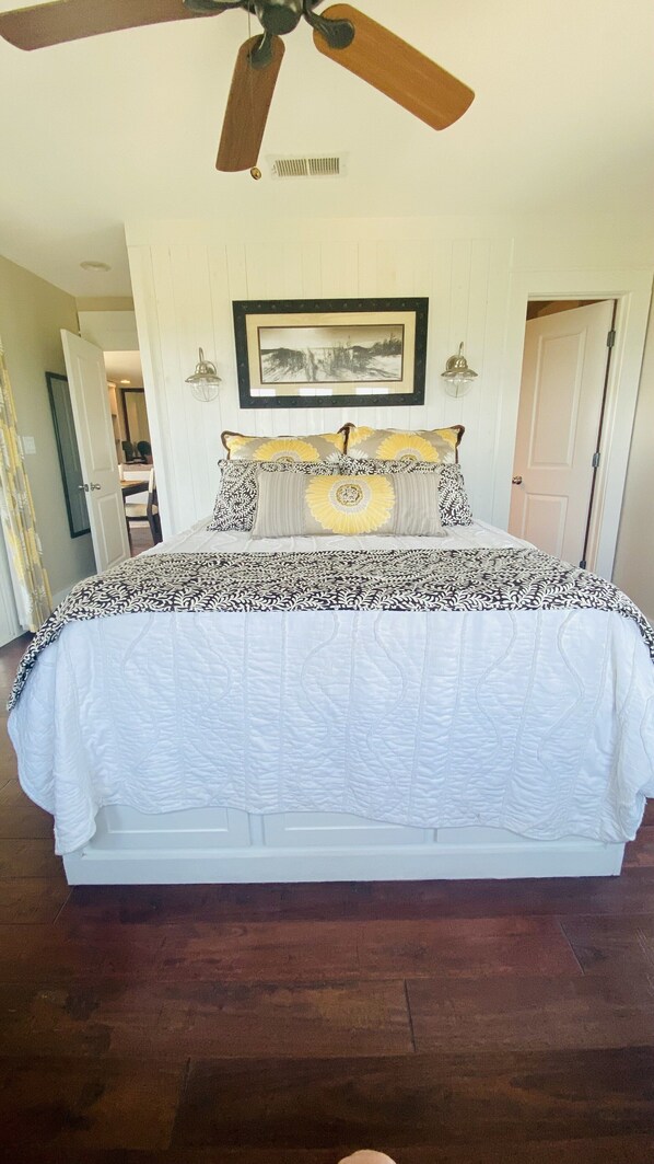 A bedroom with a large bed, a ceiling fan, a framed picture on the wall, and a door leading to another room. - OCEANFRONT LUXURY w/POOL! Professionally Decorated Perfect Beach Property 💙! (Kill Devil Hills)