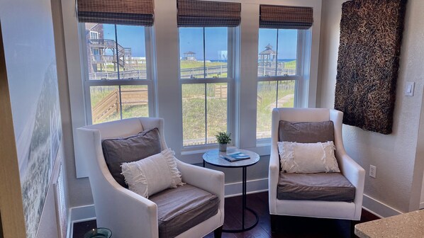 A room with two white armchairs, a small round table with a book, and a view of a beach and wooden deck through large windows. - OCEANFRONT LUXURY w/POOL! Professionally Decorated Perfect Beach Property 💙! (Kill Devil Hills)