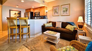 A living room with a sofa, chairs, a coffee table, and a kitchen area with wooden cabinets and appliances.