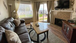 Una sala de estar con sofá de cuero, una mesa de centro, un televisor de pantalla plana montado sobre una chimenea y vistas al exterior a través de grandes ventanales.