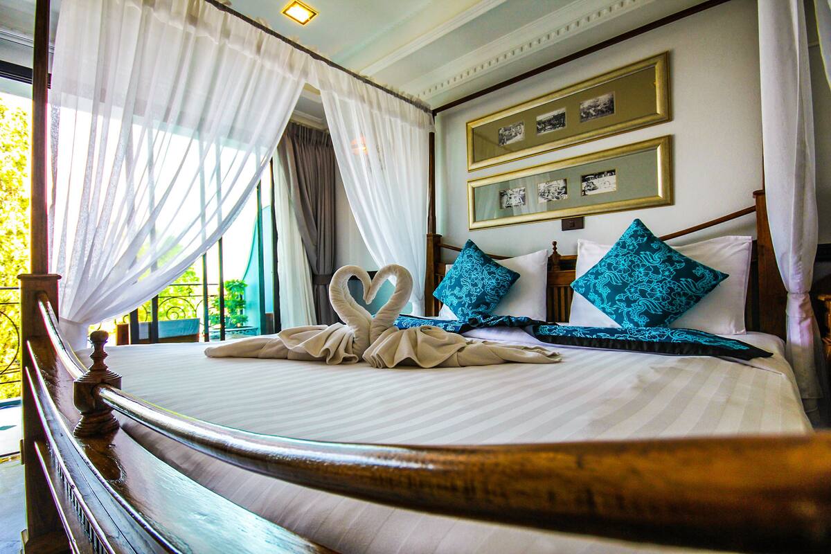 A neatly made bed with a swan-shaped towel and two blue pillows, a wooden headboard, and a view of greenery outside.