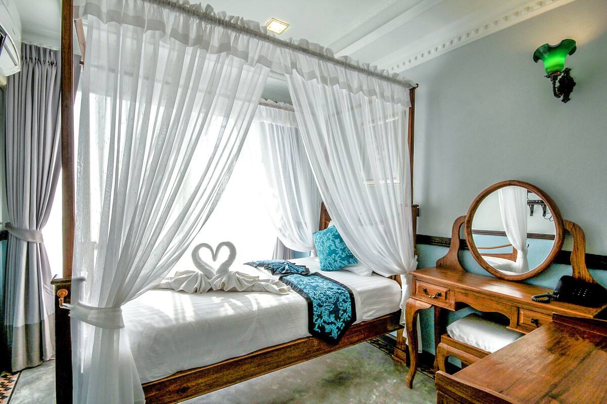 A four-poster bed with white drapes, a wooden bedside table, a mirror, and a blue pillow.