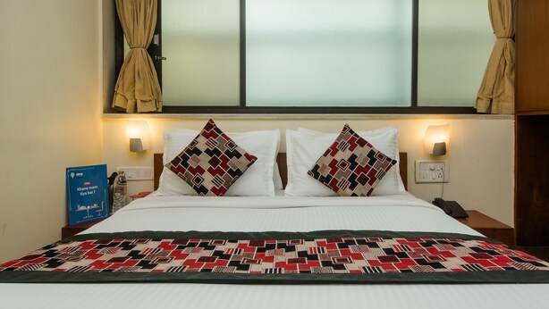 A hotel room with a bed, two pillows, a nightstand, a wall-mounted lamp, and a window with curtains. - Hotel Jayshree (Mumbai)