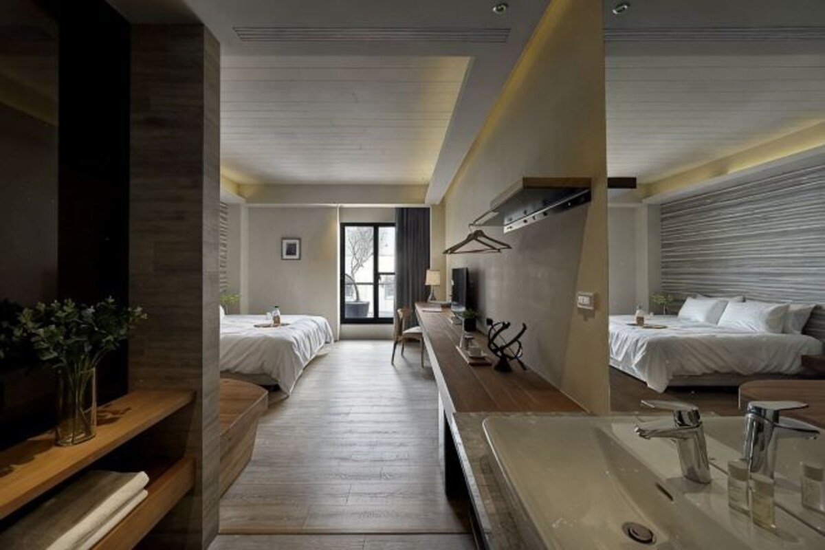A hotel room with two beds, a large window, a desk, and a bathroom with a sink and mirror.