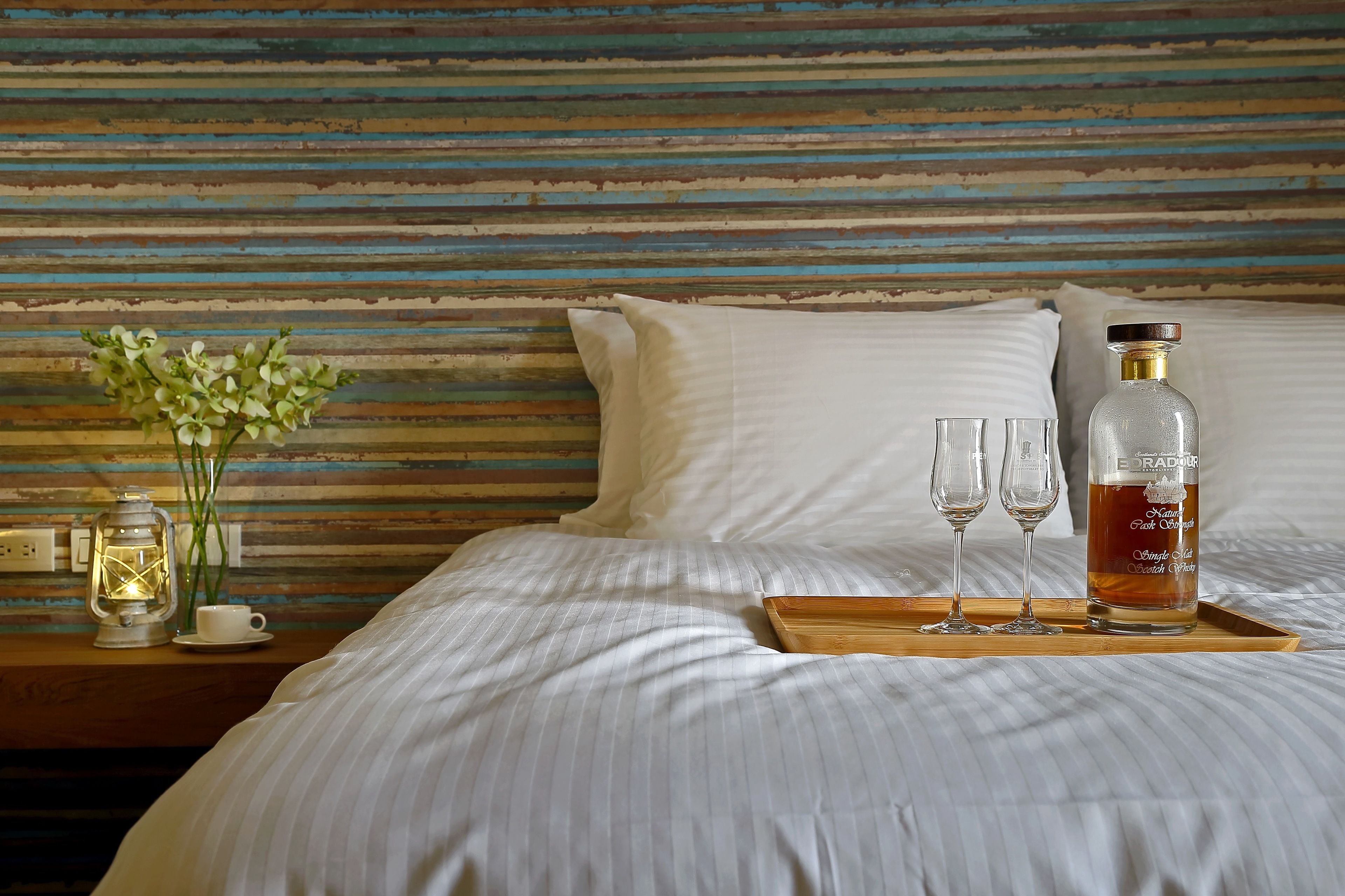 A neatly made bed with white linens, a tray with a bottle of Radisson Single Malt Scotch Whisky, two glasses, and a small vase with flowers on a wooden nightstand.