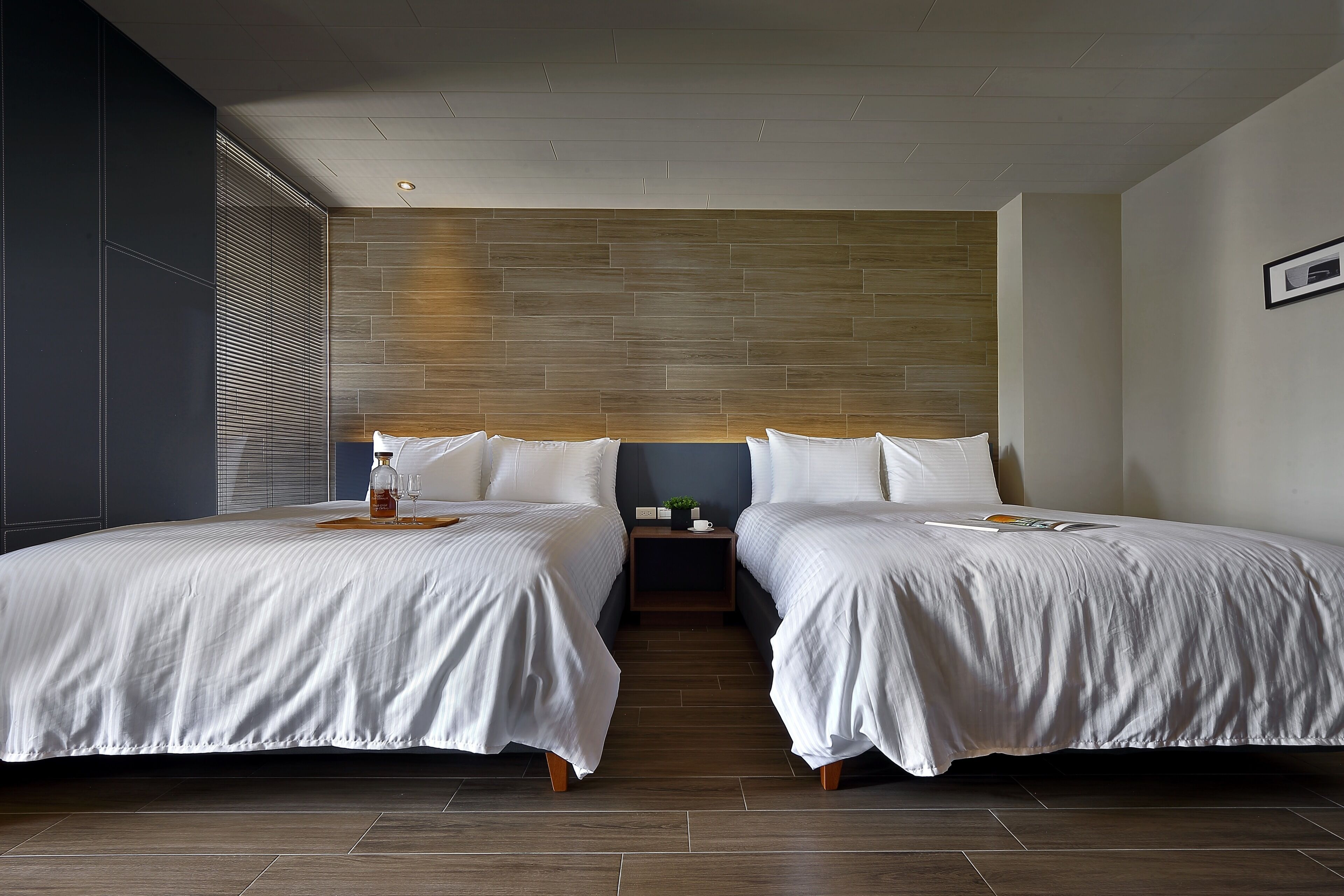 A hotel room with two beds, a wooden accent wall, and a small bedside table with a plant and a glass.
