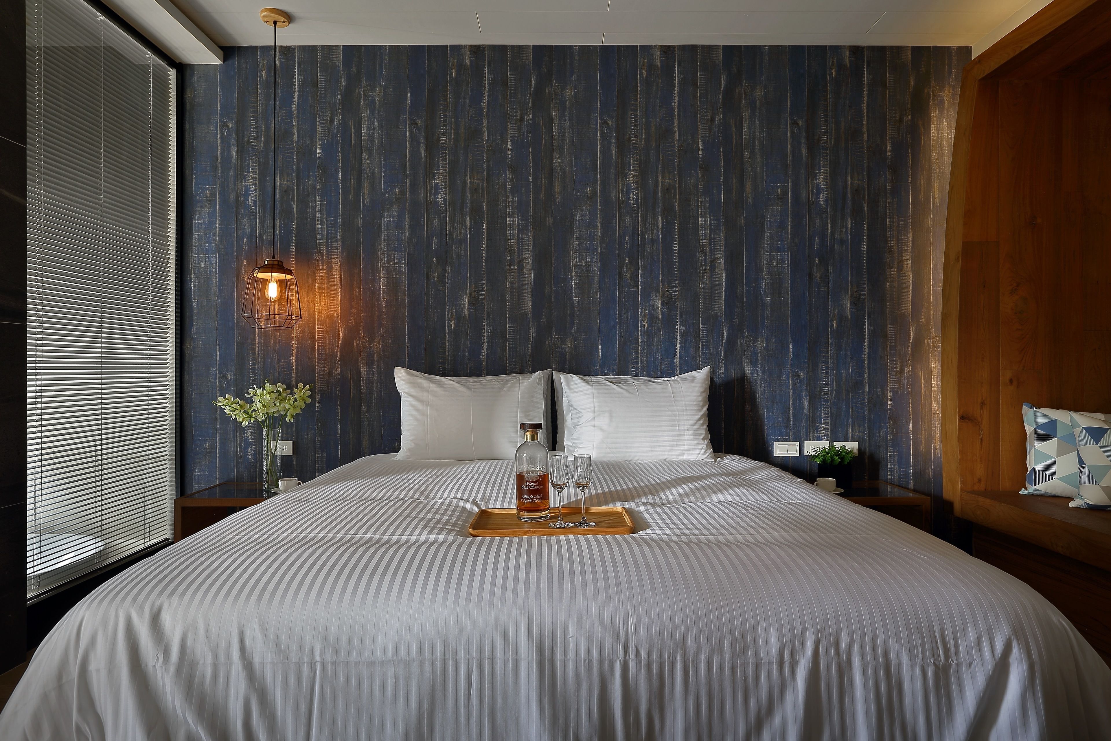 A neatly made bed with a tray of drinks and glasses, a vase with flowers, and a wall-mounted light fixture.
