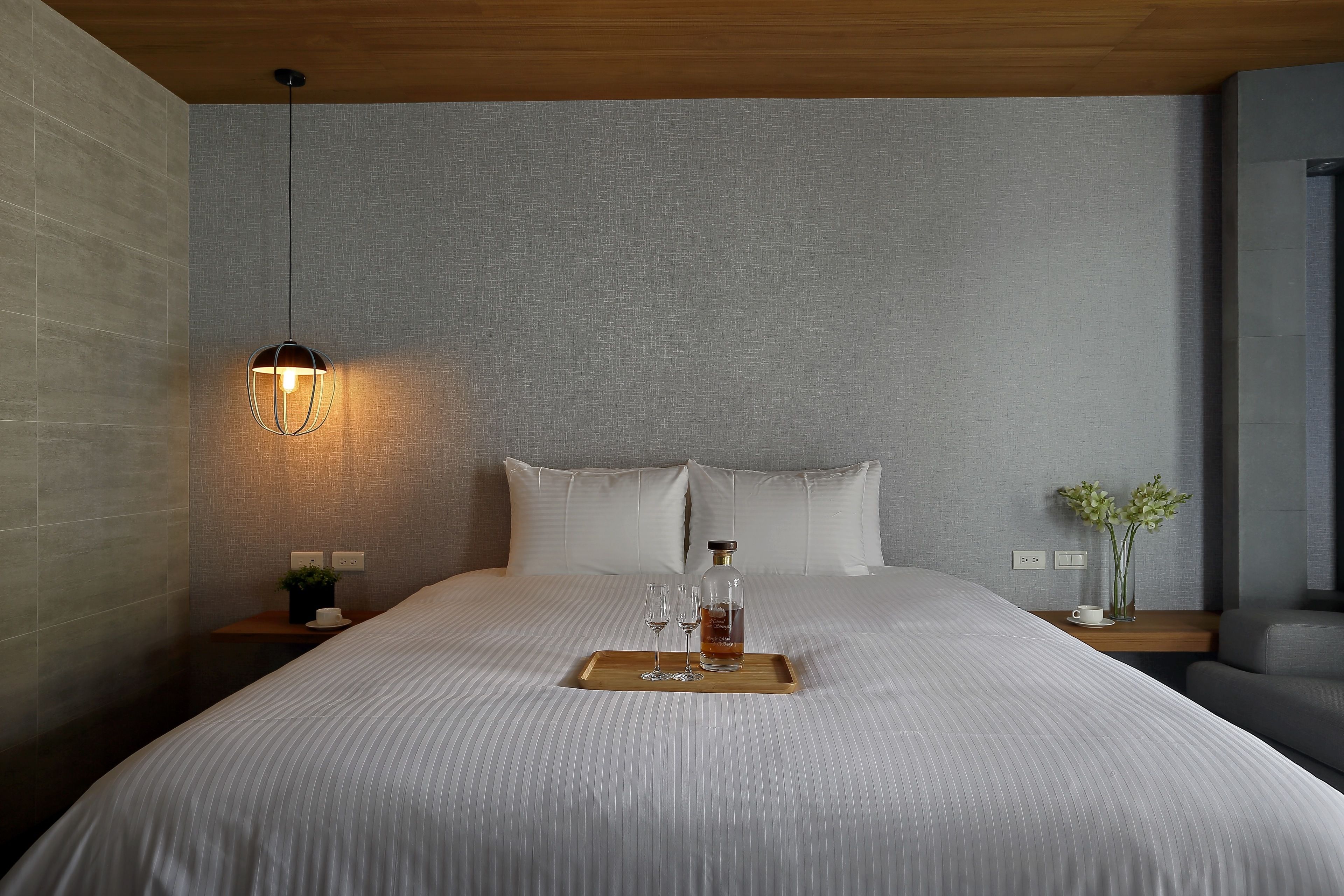 A neatly made bed with a tray holding a bottle and glasses, a bedside table with a plant, and a hanging light fixture.