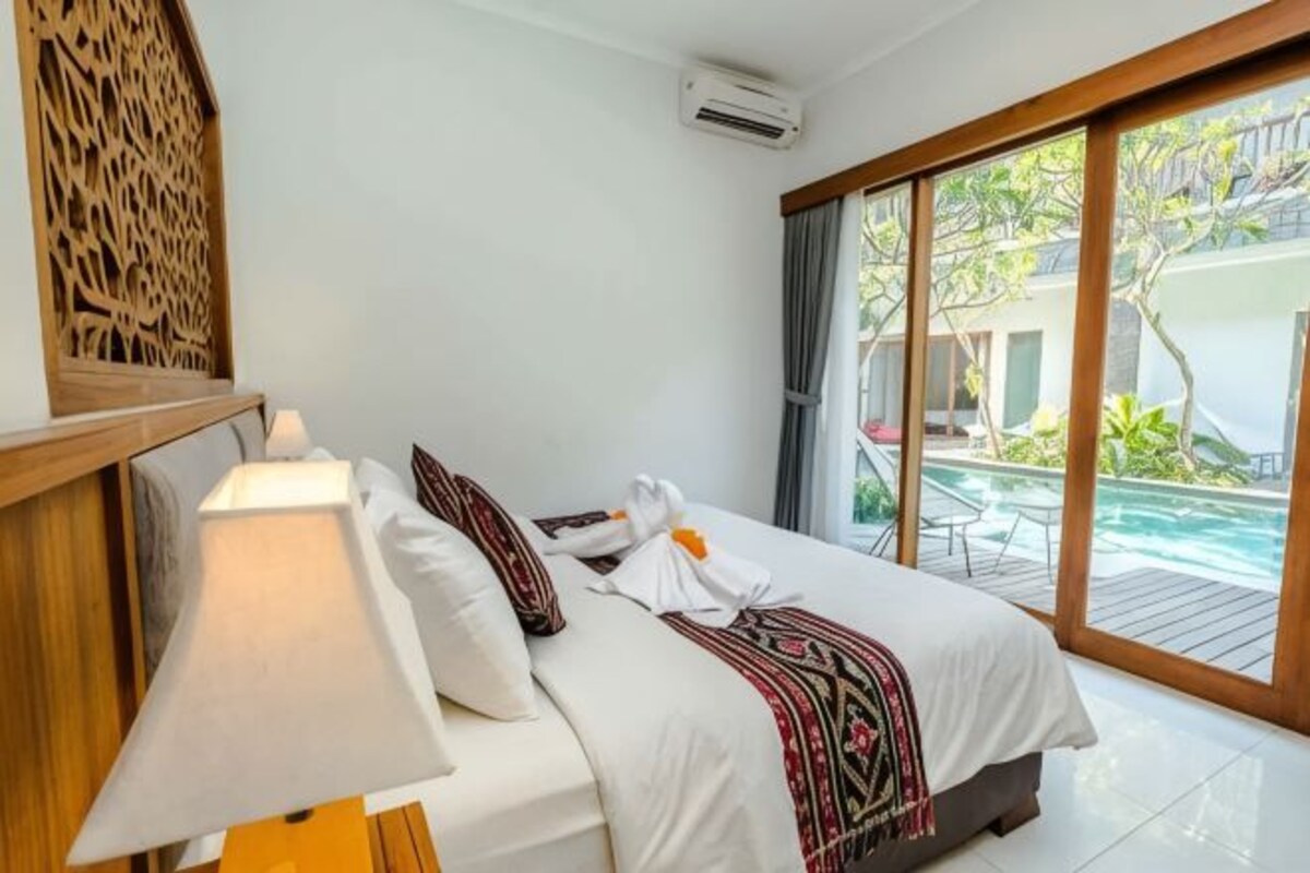 Een hotelkamer met een bed, nachtkastje, lamp en uitzicht op een buitenzwembad.