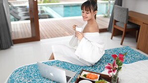 A person sitting on a bed with a laptop, holding a cup, in a room with a pool view.