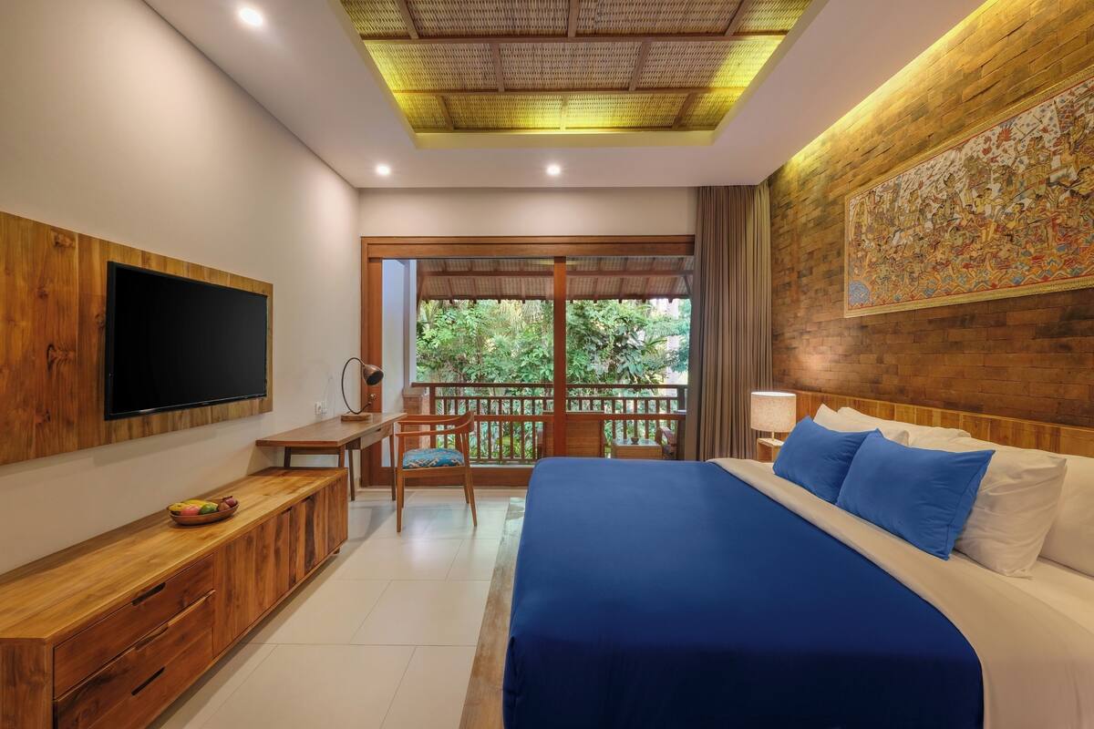 Habitación de hotel con una cama grande, un escritorio de madera, televisión y un balcón con vistas a zonas verdes.