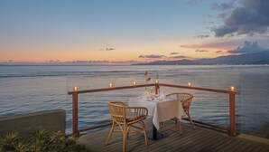 A dining area with a table and chairs overlooking a body of water at sunset. - Sagara Candidasa by AHM (Karangasem)