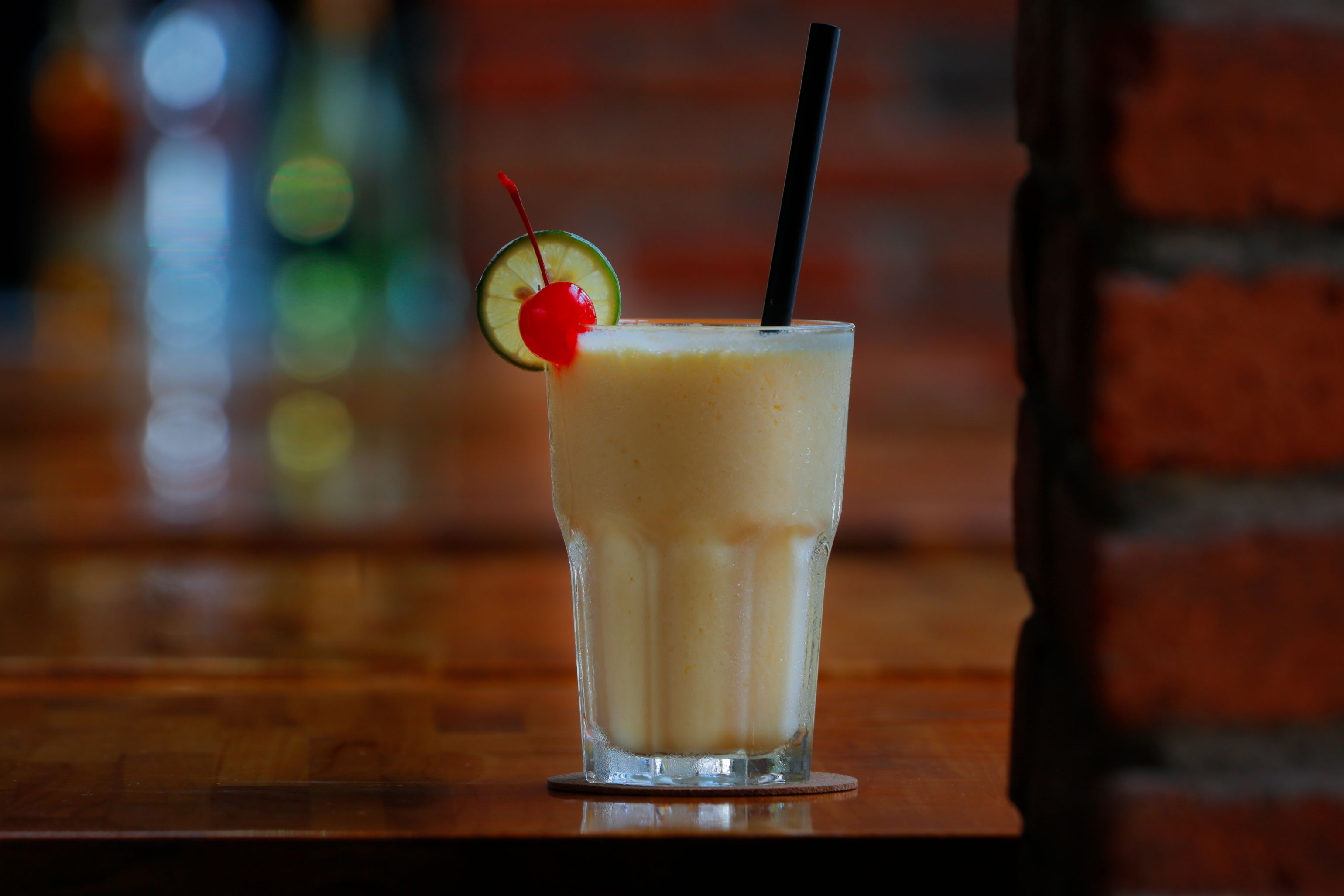 A glass of a creamy beverage with a cherry and lime garnish, a black straw, and a coaster on a wooden surface.