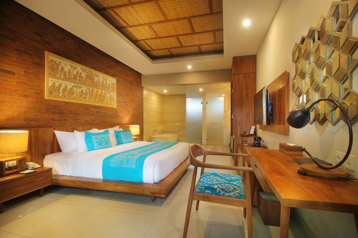 Una habitación de hotel con una cama de madera, un escritorio, una silla, una mesita de noche, una lámpara y un televisor montado en la pared.