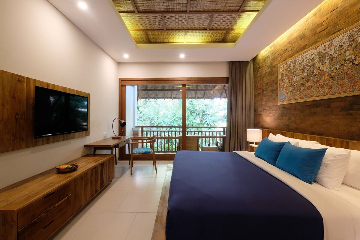 Un dormitorio con una cama grande, un escritorio, una silla, un televisor y un balcón con vistas a zonas verdes.
