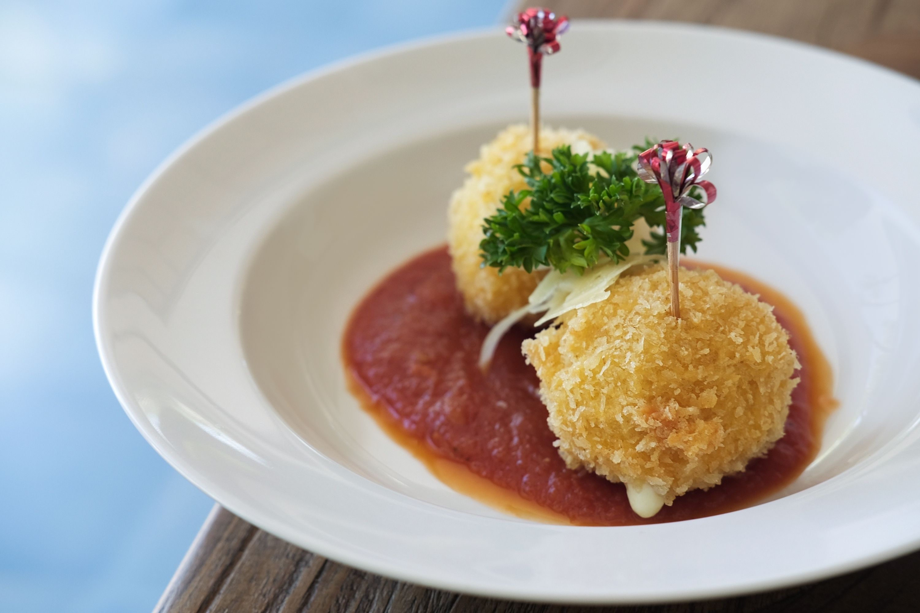 A white plate with two breaded balls of food, garnished with parsley and a decorative element, served on a wooden table.