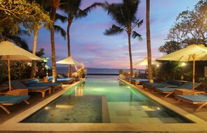 Zona junto a la piscina con sillas de descanso y sombrillas, palmeras y vista al océano al atardecer.